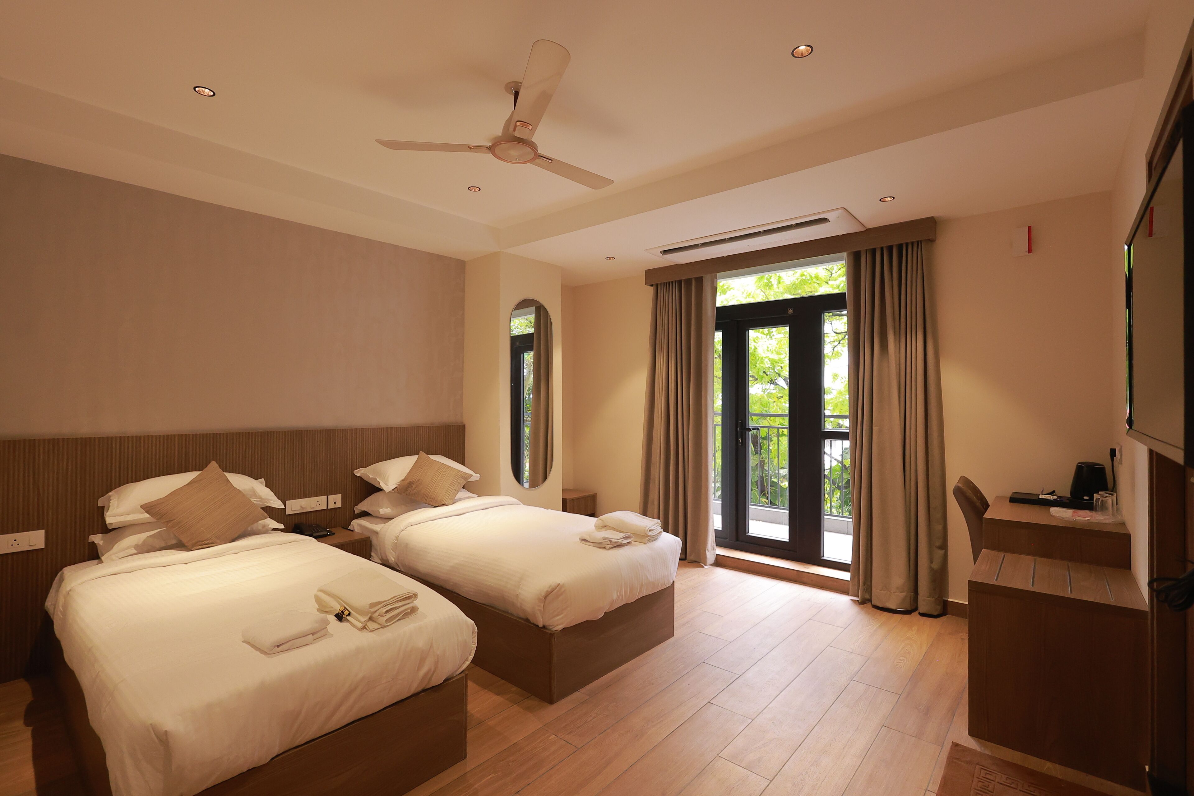 Habitación de hotel con dos camas, ventilador de techo, ventana grande con cortinas y una pequeña mesa con cafetera.