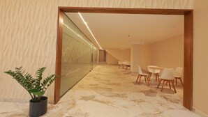 A hallway with a plant, a glass wall, and a seating area with chairs and a table.