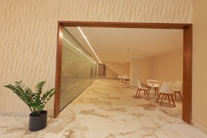 A hallway with a plant, a glass wall, and a seating area with chairs and a table.