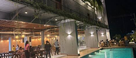 An indoor swimming pool area with a dining area adjacent to it, featuring a modern architectural design with exposed ceilings and hanging plants.