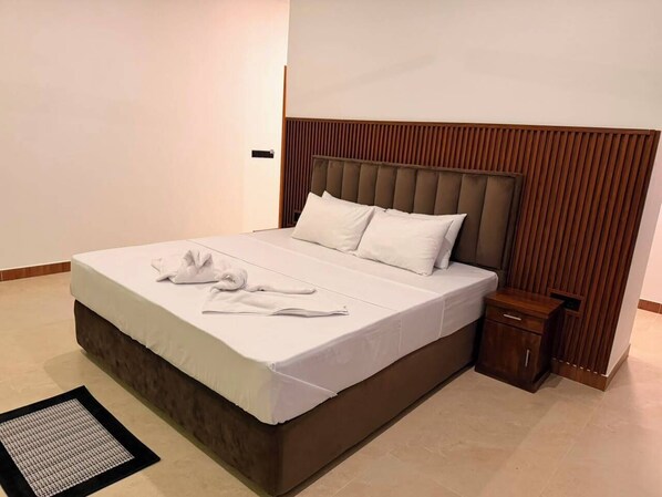 A neatly made bed with white linens and a brown headboard, a small wooden nightstand with a drawer, and a black and white area rug on a tiled floor.