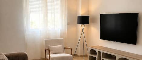 Un salon comprenant un canapé d’angle, un fauteuil, une table basse, un lampadaire et un téléviseur fixé au mur.