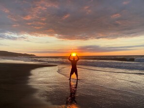 A beach scene with a person standing on the wet sand, facing the sunset.