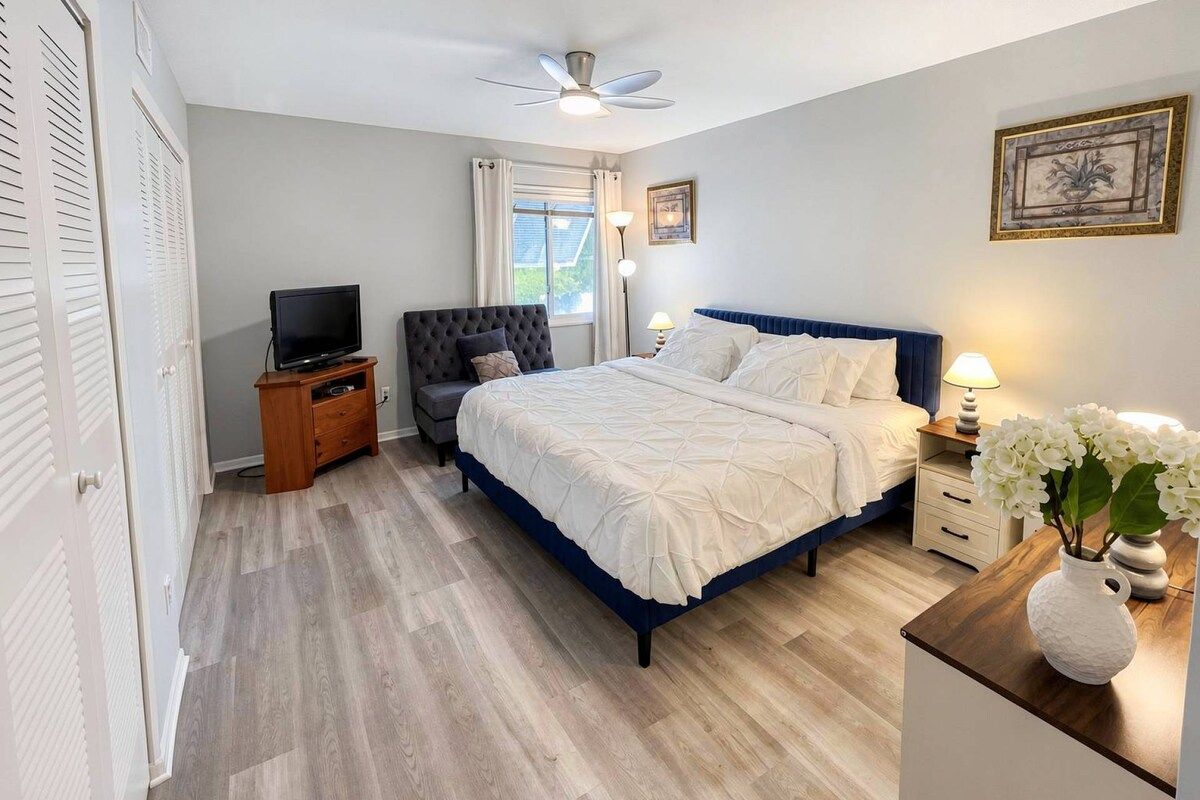 Une chambre à coucher comprenant un lit, des tables de chevet, un téléviseur, un ventilateur de plafond et une fenêtre avec des rideaux.
