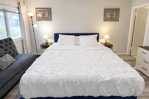 A bedroom with a large bed, two bedside tables with lamps, a grey sofa, and two wall-mounted pictures. - Renovated Condo Across from Siesta Key Beach | (Siesta Key)