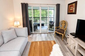 Ein Wohnzimmer mit einer Couch, einem Sessel, einem Fernseher, einem Holztisch und einer Glastür, die zu einem Außenbereich mit Möbeln führt.