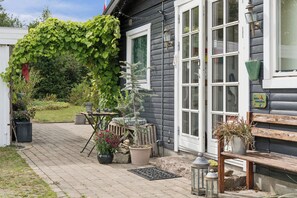 A house with a dark exterior, a white door, and a window with a white frame. There is a small tree in front of the house, and various potted plants and a metal lantern on the porch. - Cozy house by Klitgård Fishing Village (Nibe)