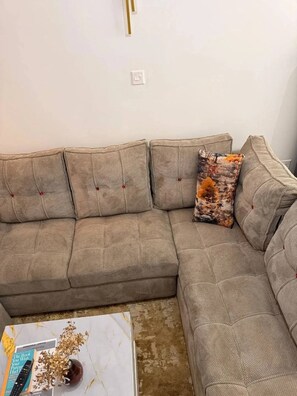 Ein beiges Ecksofa mit einem gemusterten Zierkissen, ein Couchtisch mit einem Buch und dekorativen Gegenständen sowie eine weiße Wand mit einem Lichtschalter.