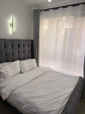 A bedroom with a large bed, a headboard, and a window with sheer curtains. - Comfort homes with free wifi  (Nairobi)