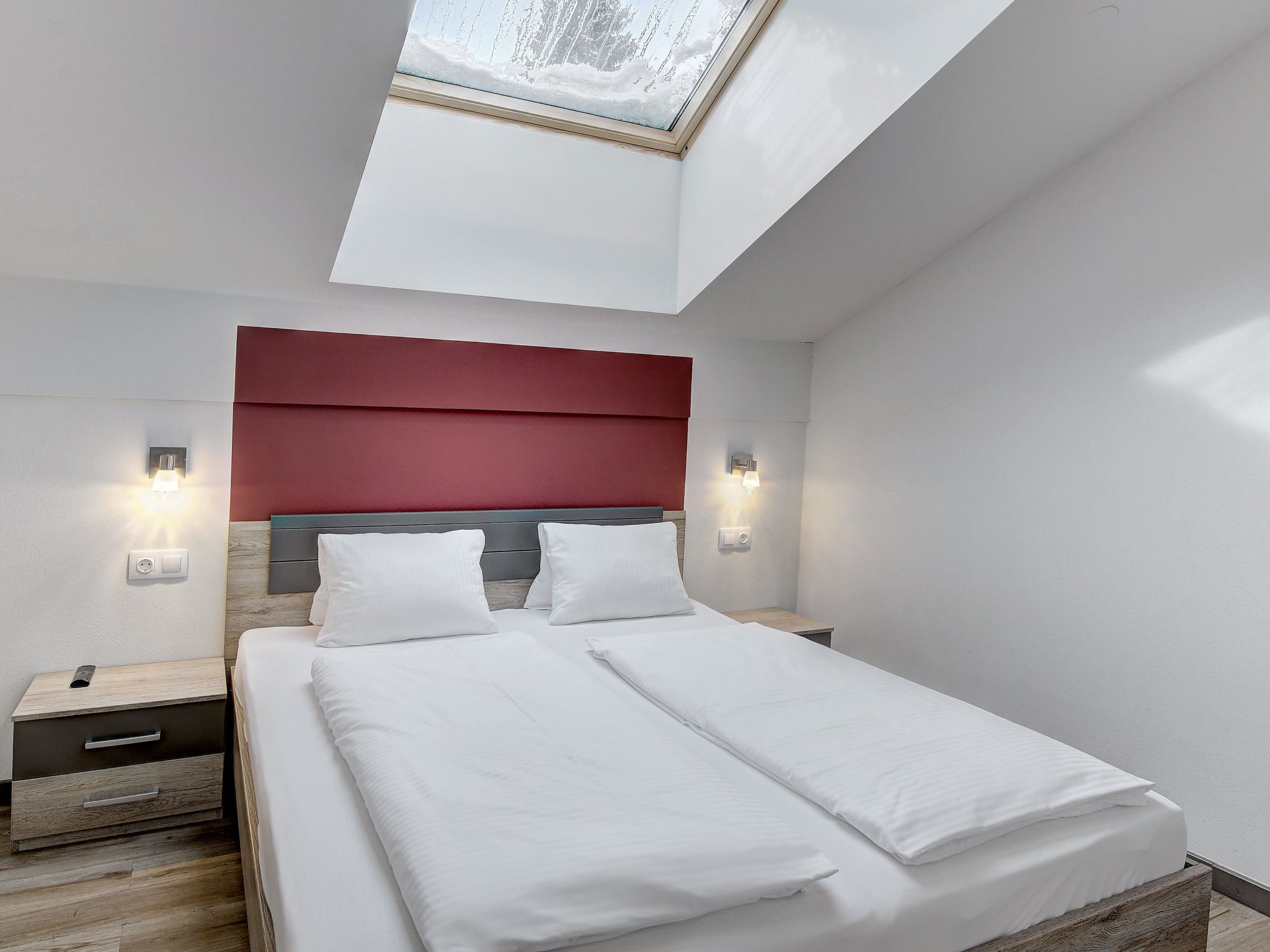 Een moderne slaapkamer met dakraam, een bed met witte lakens, een rode hoofdbord en bedlampjes.