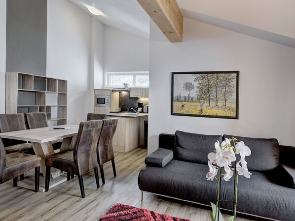 En moderne stue med en grå sofa, et spiseområde med et træbord og stole, samt et køkken med mikroovn og kaffemaskine.