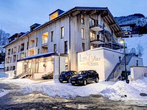 A multi-story hotel with a wooden facade, a covered entrance, and a parking area with cars. - LoriVita by Interhome (Saalbach-Hinterglemm)