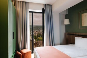 Habitación de hotel con cama, ventana con cortinas, vista a un pueblo y una lámpara fijada a la pared.