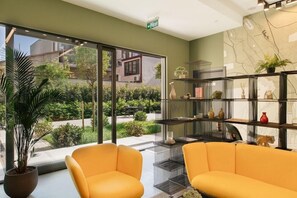 A room with two yellow chairs, a large window with a view of greenery and buildings, and a black metal shelf with decorative items. - Hotel 21, Tbilisi (Tbilisi)
