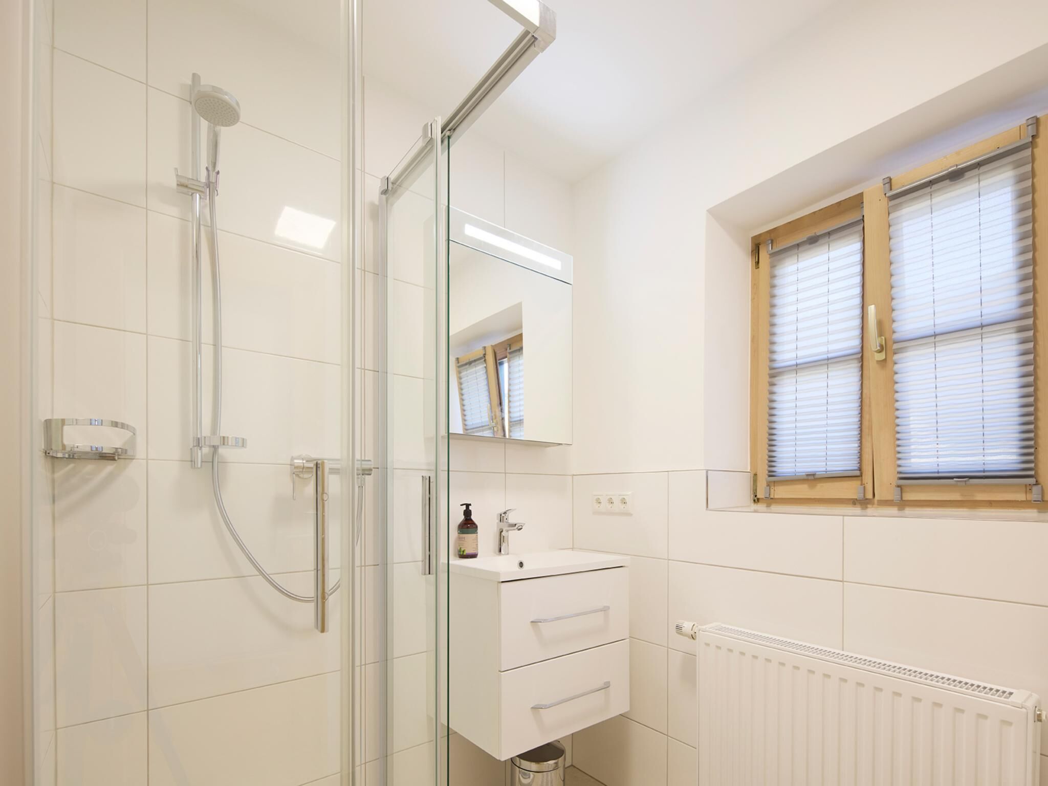 A bathroom with a glass shower enclosure, a white sink with a mirror above it, and a window with blinds.