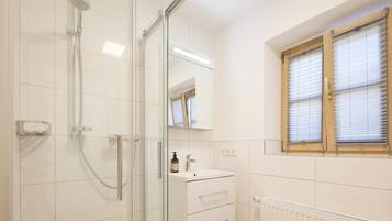 A bathroom with a glass shower enclosure, a white sink with a mirror above it, and a window with blinds.