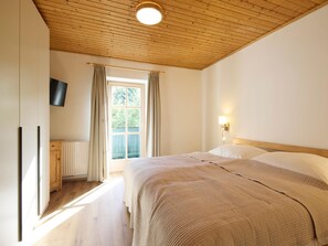 A bedroom with a wooden ceiling, a bed with a beige blanket, a window with curtains, and a wall-mounted TV.