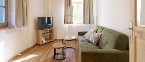 A room with a wooden ceiling, a television on a wooden stand, a green sofa, a small round table, and a window with curtains.