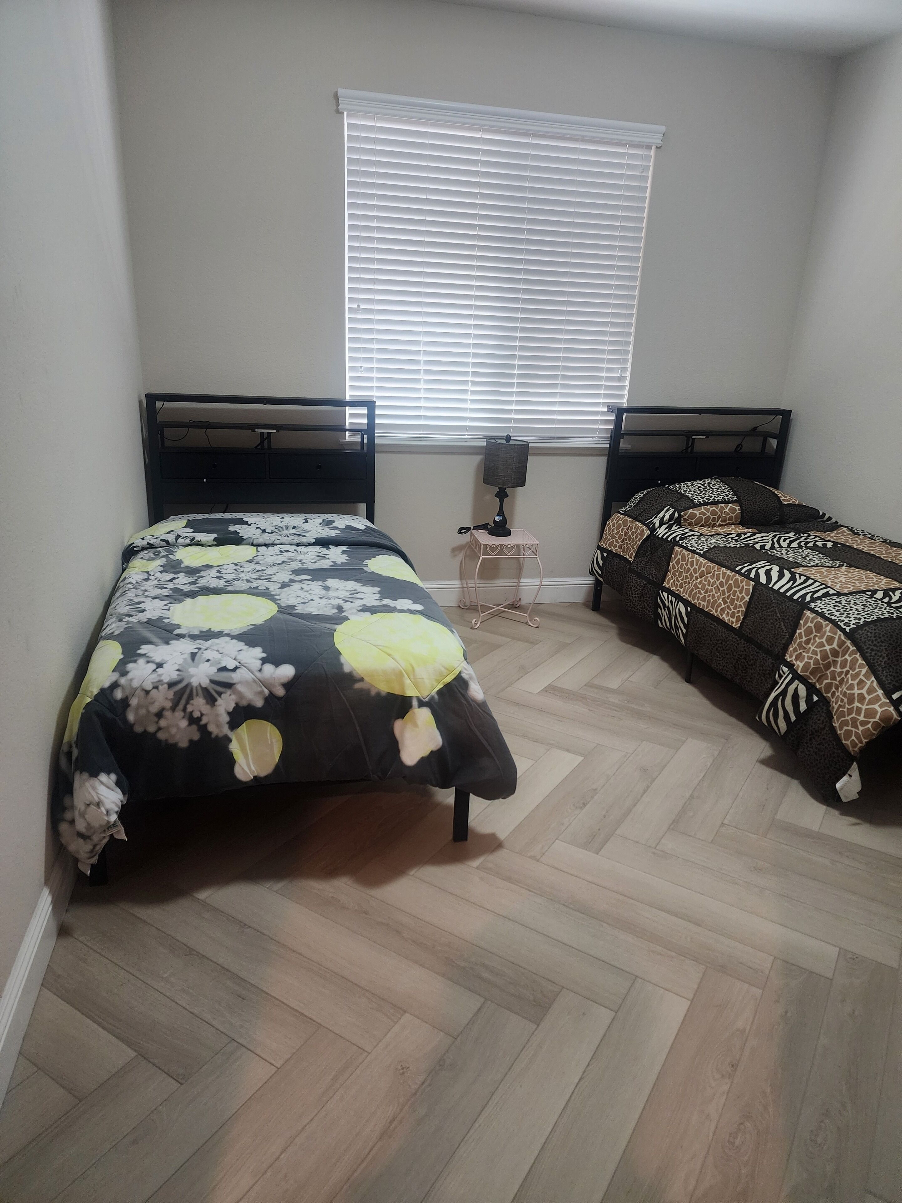 A bedroom with two beds, a window with blinds, a bedside table with a lamp, and a herringbone-patterned floor.