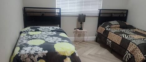 A bedroom with two beds, a window with blinds, a bedside table with a lamp, and a herringbone-patterned floor.