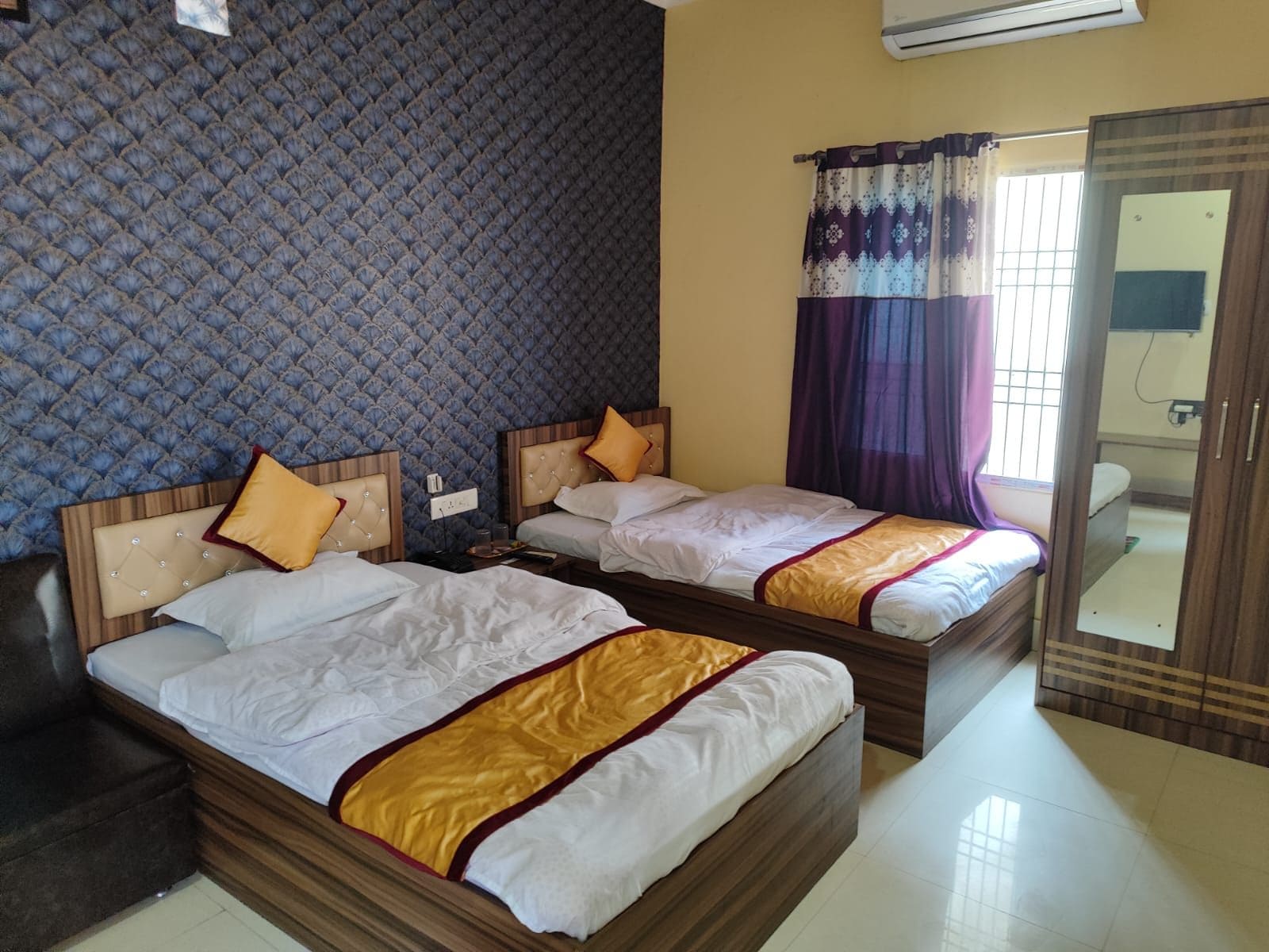 A hotel room with two beds, a patterned wall, a window with curtains, and a mirror.