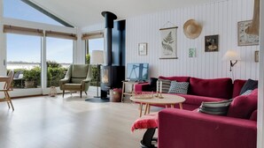 A modern living room with a large window, a fireplace, a red sofa, and a television. - Stunning Sea Views, Wood-Fired Hot Tub & Nature (Haarby)
