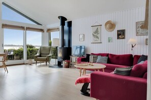 A modern living room with a large window, a fireplace, a red sofa, and a television. - Stunning Sea Views, Wood-Fired Hot Tub & Nature (Haarby)