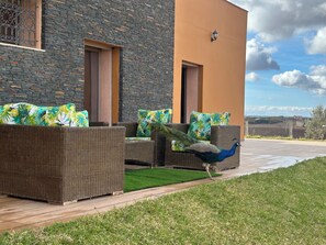 A peacock on a patio with wicker furniture and a stone wall.