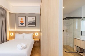 Ein Hotelzimmer mit einem Bett, zwei Wandbildern, einem Nachttisch mit Lampe sowie einem modernen Badezimmer mit Duschkabine aus Glas.