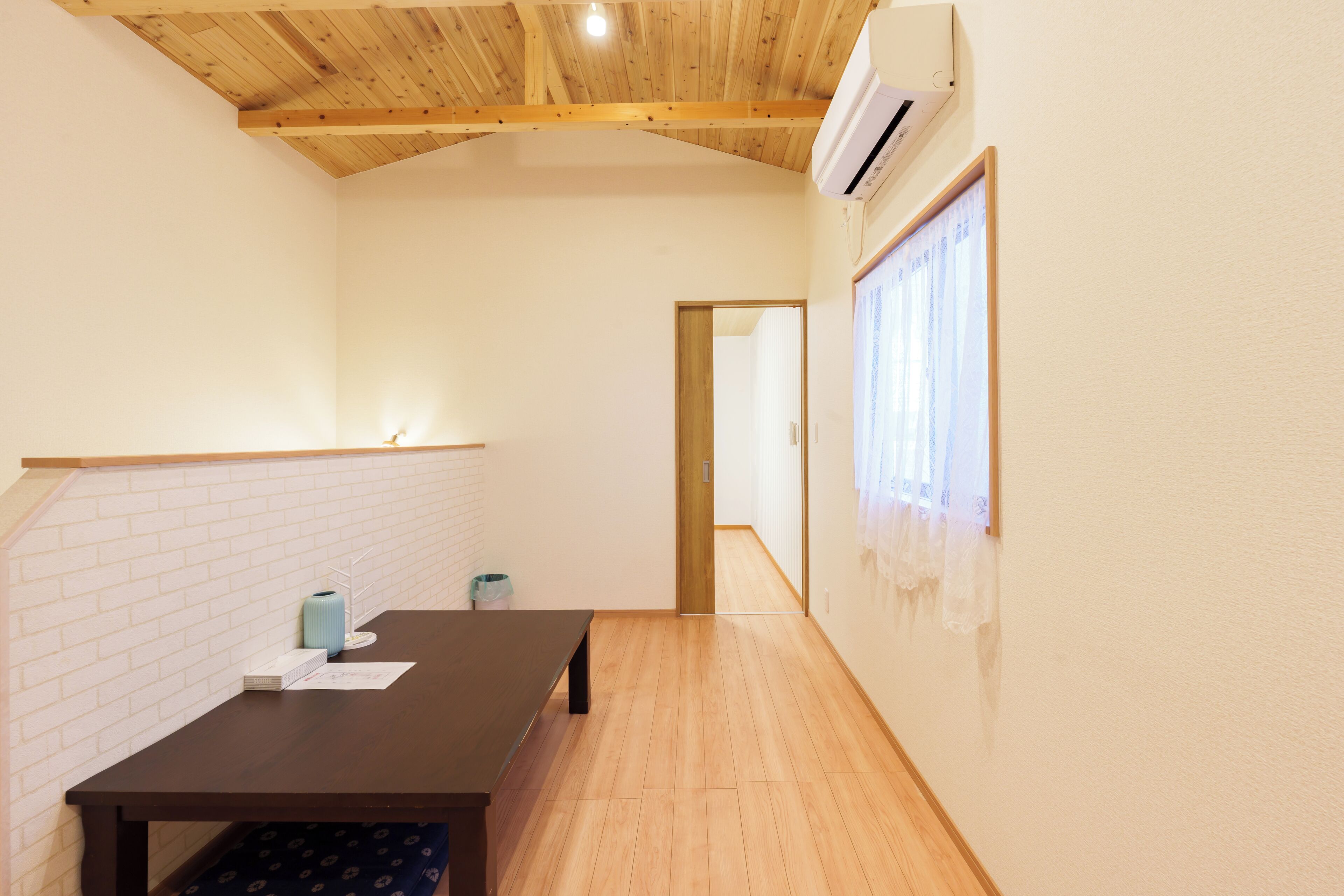 A room with a wooden ceiling, a table with papers, a trash bin, and a window with curtains.