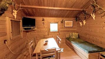 A cabin interior with a wooden dining table, a TV mounted on the wall, a window, and a bed with a green mattress.