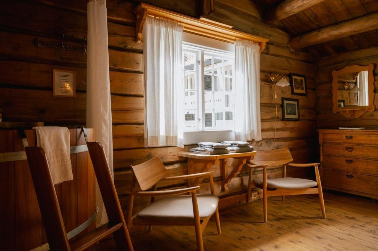 A wooden cabin interior with a dining table, two chairs, a window with curtains, a dresser, and a mirror.