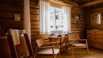 A wooden cabin interior with a dining table, two chairs, a window with curtains, a dresser, and a mirror.