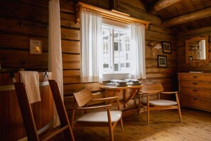 En trästuga med en matplats, ett bord och två stolar, ett fönster med gardiner, en byrå och en spegel.