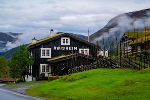 A black building with 'ROISHEIM' written on the front, surrounded by a grassy area and mountains in the background. - Roisheim Hotell (Lom)