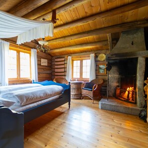 Interior de cabana de madeira com cama, lareira e uma poltrona de vime.