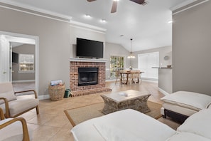 A spacious living room with a fireplace, a television mounted above it, and a dining area with a table and chairs. - Ocean Springs Bayou Getaway | Outdoor Living (Gulf Park Estates)