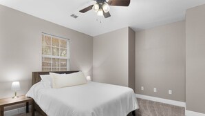 A bedroom with a bed, nightstand, lamp, window with blinds, and a ceiling fan. - Ocean Springs Bayou Getaway | Outdoor Living (Gulf Park Estates)