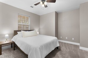Ein Schlafzimmer mit Bett, Nachttisch, Lampe, Fenster mit Jalousien und Deckenventilator.