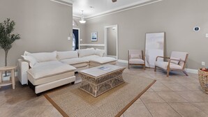 A living room with a sectional sofa, a coffee table, two armchairs, a ceiling fan, and a door leading to another room. - Ocean Springs Bayou Getaway | Outdoor Living (Gulf Park Estates)