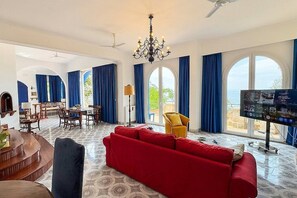 Amplia sala de estar con un sofá rojo, un sillón amarillo y un televisor. Cuenta con ventanas grandes con cortinas azules, un candelabro y un comedor con mesa y sillas.