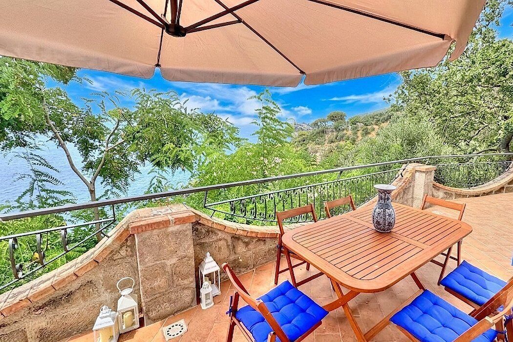 An outdoor patio with a wooden table and blue cushioned chairs, a beige umbrella, and a view of a natural landscape.