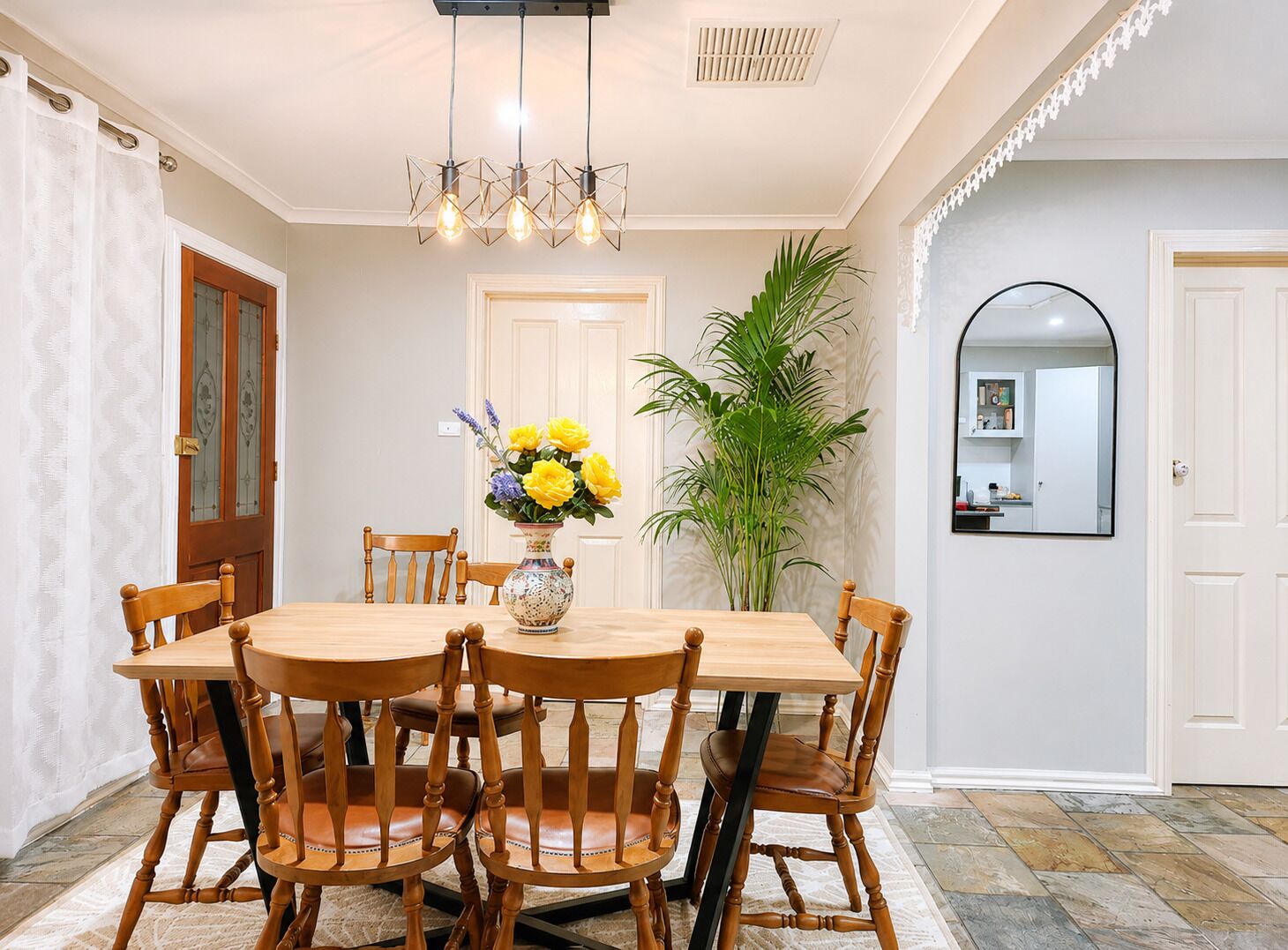 A dining area with a wooden table and chairs, a vase with flowers, and a large plant.