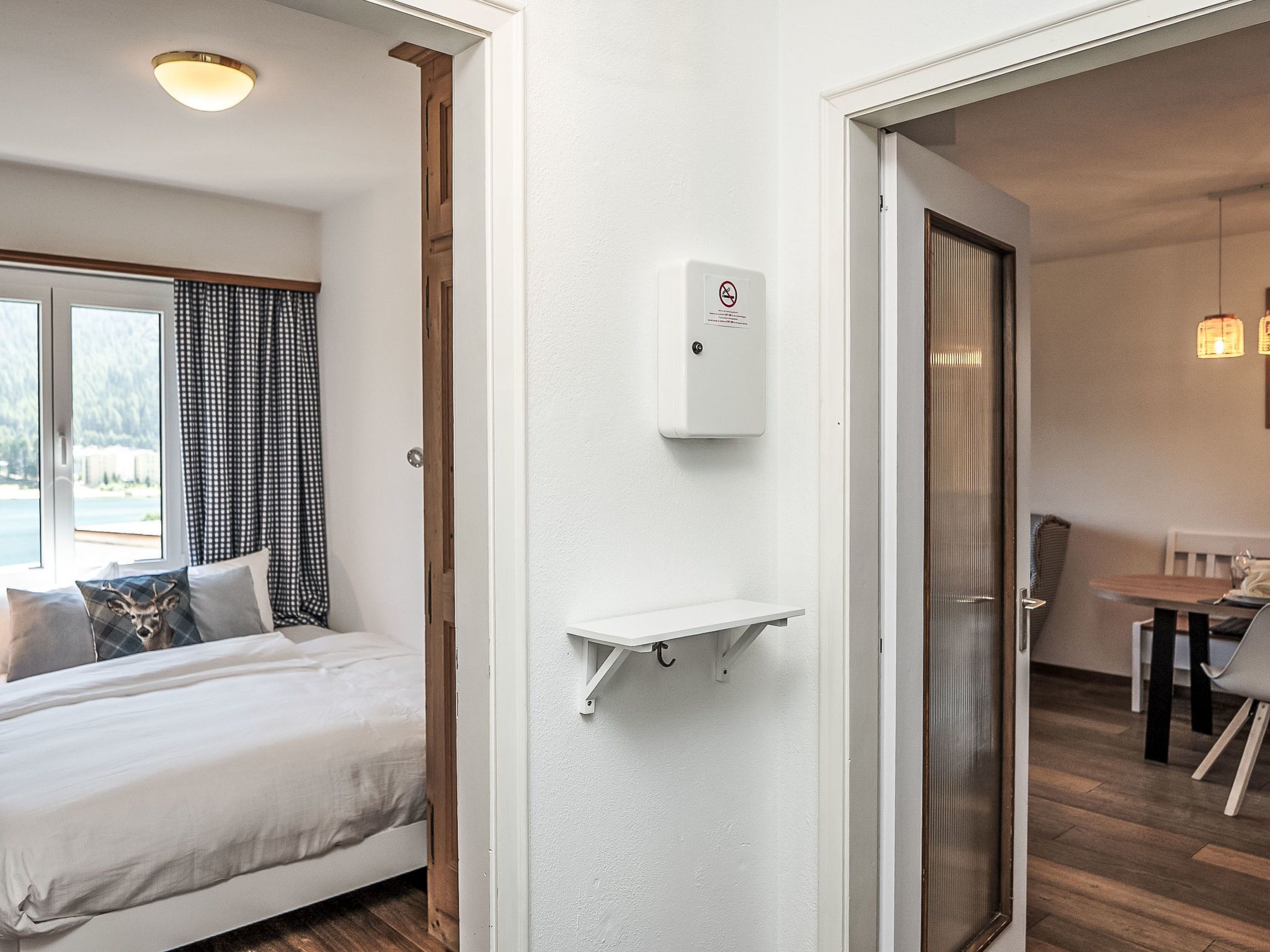 A hotel room with a bed, a small white shelf, a door leading to a dining area, and a window with curtains.