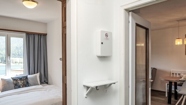Hotellihuone, jossa on sänky, pieni valkoinen hylly, ovi, joka johtaa ruokailutilaan, ja ikkuna, jossa on verhot.