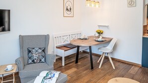 Moderni olohuone, jossa on ruutukuviollinen nojatuoli, puinen ruokapöytä valkoisine tuoleineen, penkki ja pieni sivupöytä, jonka päällä on kuppi.