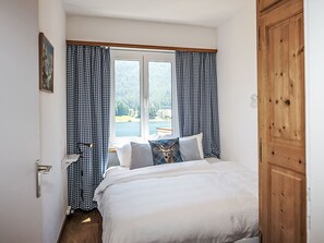 Ein Schlafzimmer mit einem Bett, karierten Vorhängen, einem Fenster mit Blick auf einen See und Wald sowie einer Holztür.