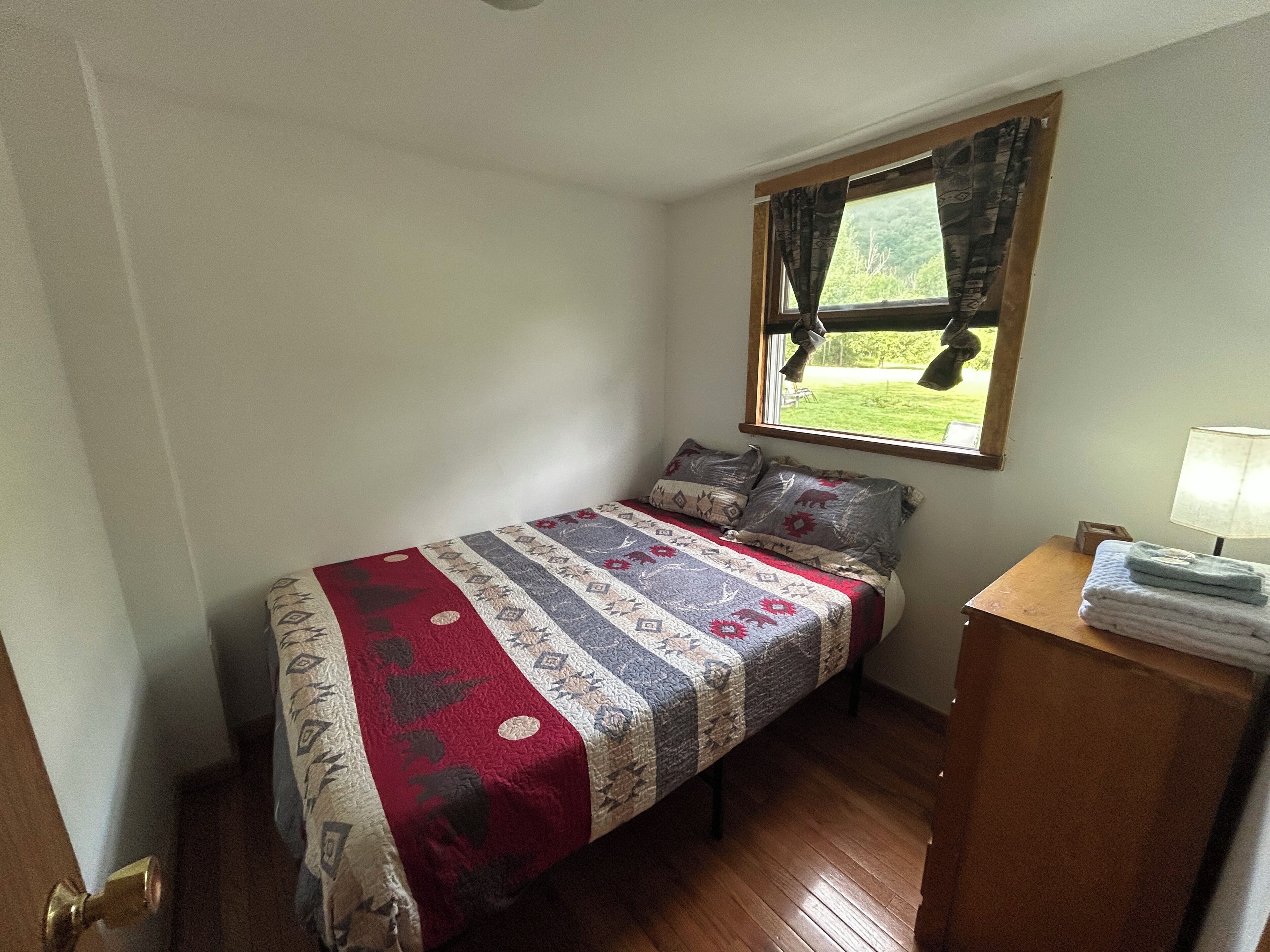 A small, single-bed room with a window, a bedside table with a lamp, and a door.