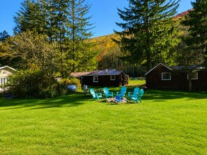Eine grüne Rasenfläche mit einer Holzhütte, einem Grill und mehreren Adirondack-Stühlen.