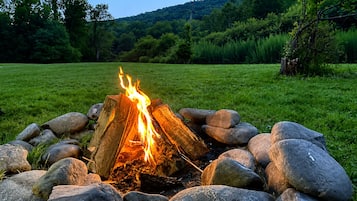 A campfire surrounded by rocks in a grassy field with trees and hills in the background.